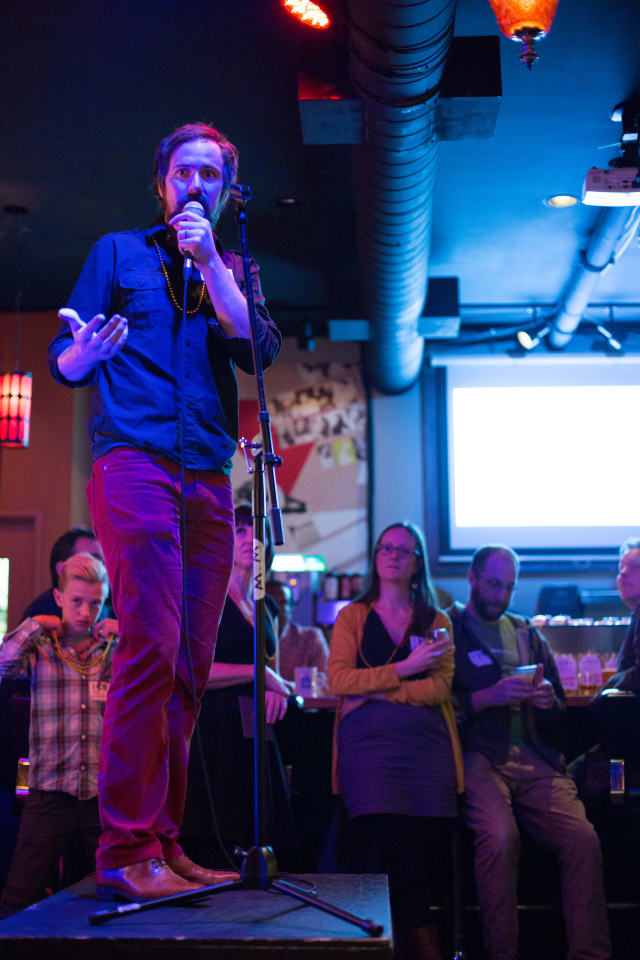 Man speaking into a microphone on stage at the Love Bomb 2017 event, with audience members in the background, showcasing community engagement and support for My Voice Music.